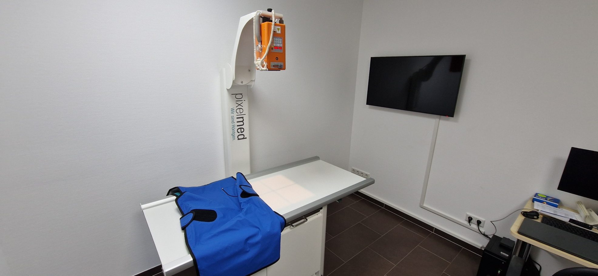 Ein Behandlungsraum mit einem Tisch, Röntgengerät und einem Monitor an der Wand.